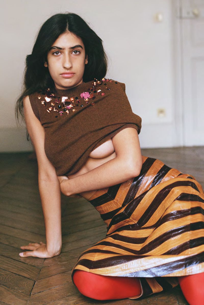 Young woman with dark hair and red tights poses in a striped skirt and brown top in an indoor setting with hardwood floors.