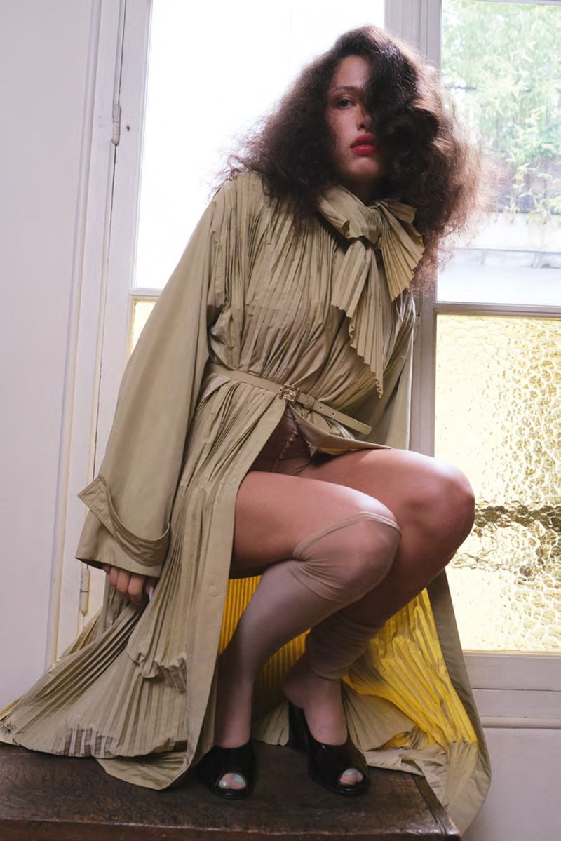 Woman in olive-green pleated trench coat and stockings poses kneeling near a window for a fashion shot with natural lighting.