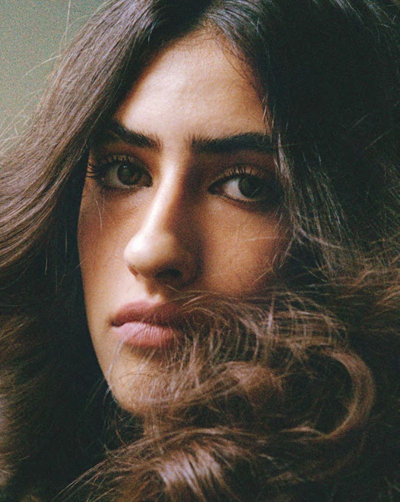 Close-up portrait of a young woman with dark wavy hair partially covering her face, looking directly into the camera.