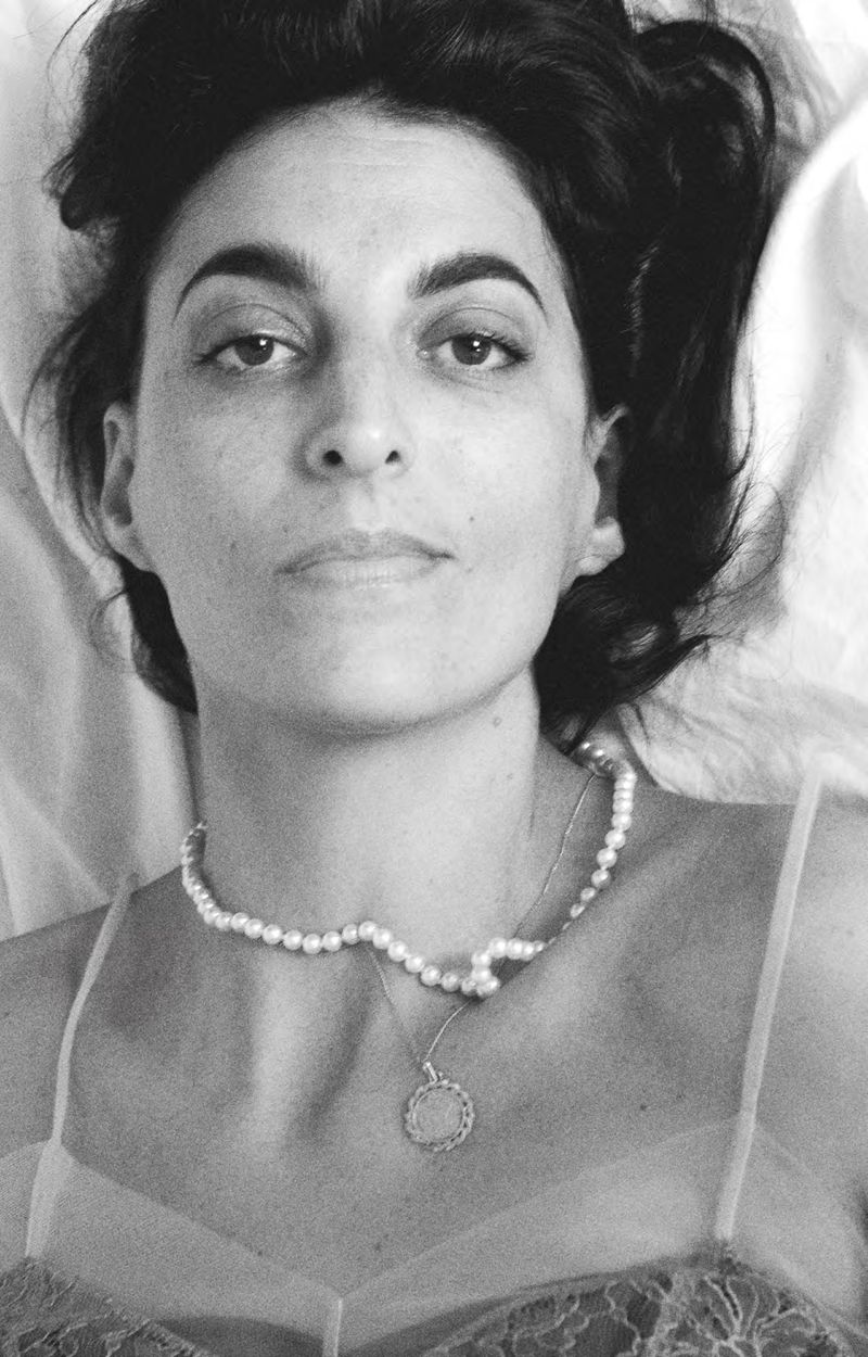 Monochrome portrait of an adult woman with dark hair laying on white sheets, wearing a pearl necklace and low-cut lacy top.