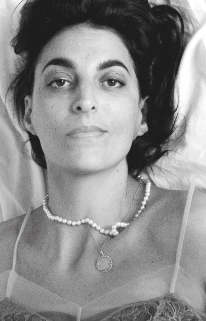 Monochrome portrait of an adult woman with dark hair laying on white sheets, wearing a pearl necklace and low-cut lacy top.