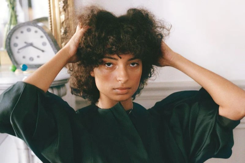 Portrait of a young adult woman with curly hair, holding her hands in her hair, wearing a dark green shirt, in an indoor setting.