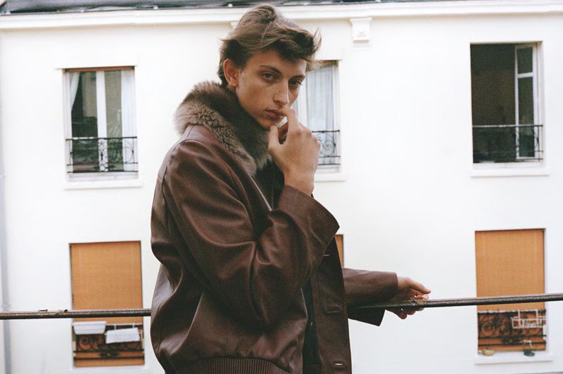 Young adult male in leather jacket with fur collar poses on balcony in front of building with windows on a cloudy day.