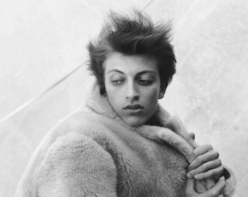 Black and white portrait of a model with textured hair wearing a plush shearling coat against a light backdrop.