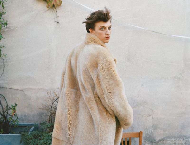 Young adult in a beige fur coat glances over their shoulder against a neutral backdrop with outdoor vegetation and a wooden chair.