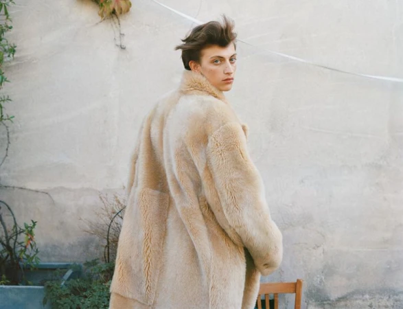 Young adult in a beige fur coat glances over their shoulder against a neutral backdrop with outdoor vegetation and a wooden chair.
