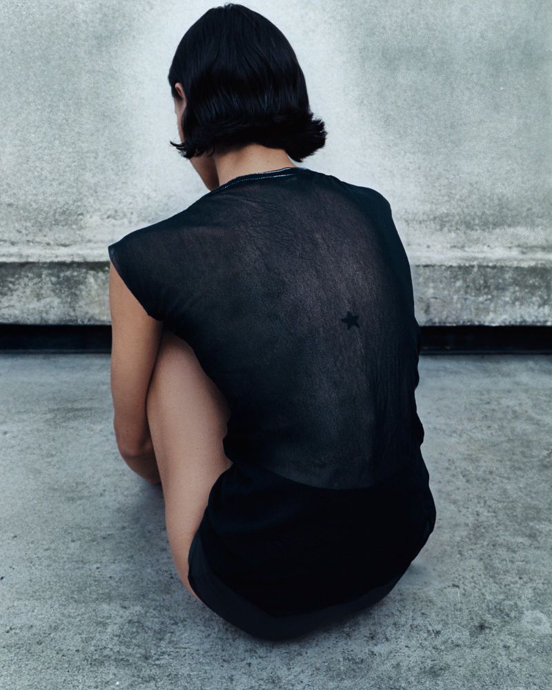Back view of a young adult woman with short black hair crouching on a gray surface wearing a sheer black top with a star detail.
