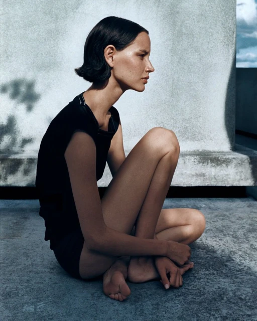 Profile portrait of a young adult woman with short dark hair sitting in shadow, wearing a black top, and against a concrete background.