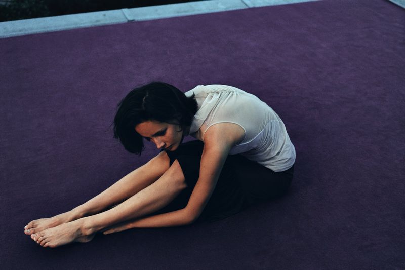 Young adult woman with dark hair sits on a purple surface, wearing a white top and dark pants, appearing thoughtful in an indoor setting.