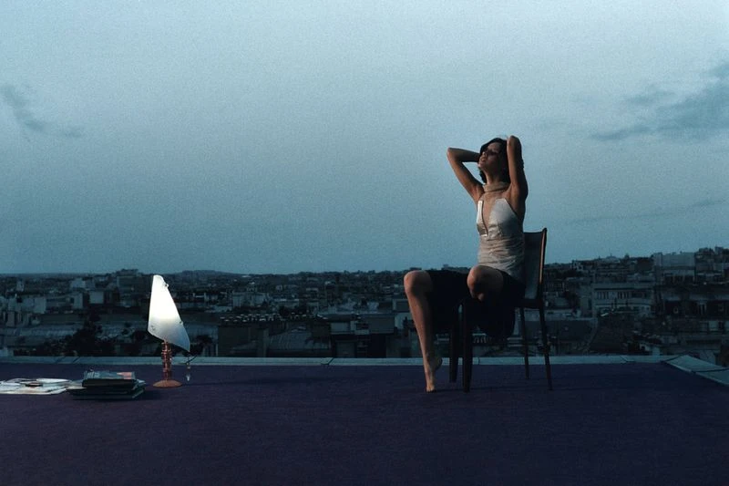 Woman sits on chair on a rooftop at dusk overlooking a city, illuminated by a lamp. She wears a light-colored dress and has her hands behind her head.