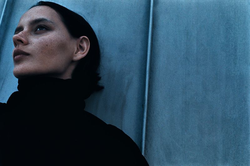 Close-up portrait of a woman with freckles wearing a black turtleneck looking upward against a cool blue wall.
