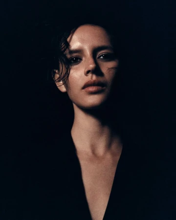 Close-up portrait of an adult female with short, dark hair, wearing a black top, set against a stark black background with dramatic lighting.