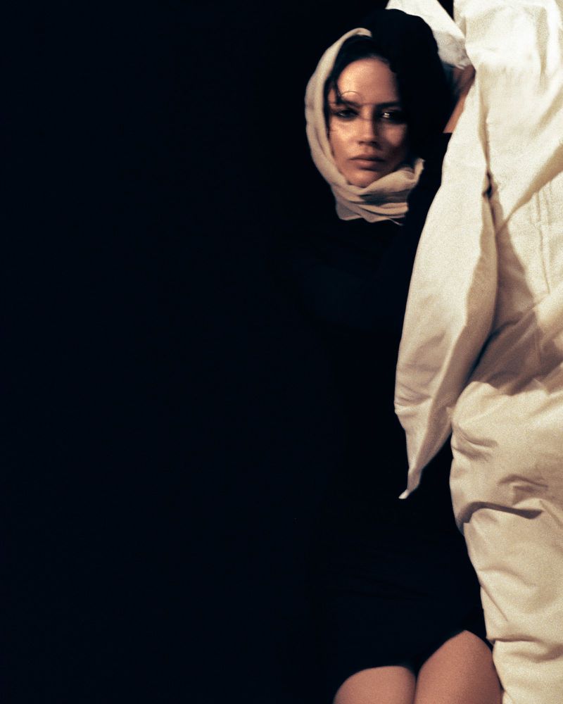 Portrait of a woman wearing a headscarf, gazing forward from a dark background, with a white robe-like garment partially in the frame.