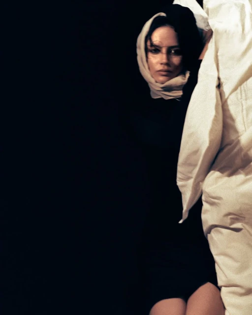 Portrait of a woman wearing a headscarf, gazing forward from a dark background, with a white robe-like garment partially in the frame.