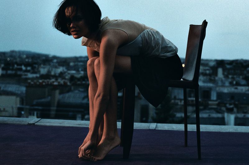Model in a light top and dark skirt seated on a chair on an open rooftop overlooking a city skyline at dusk.