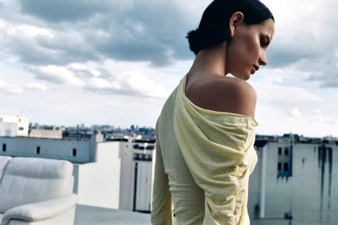 Model seen from behind wearing a draped pale yellow off-the-shoulder top against an urban cityscape from a rooftop.
