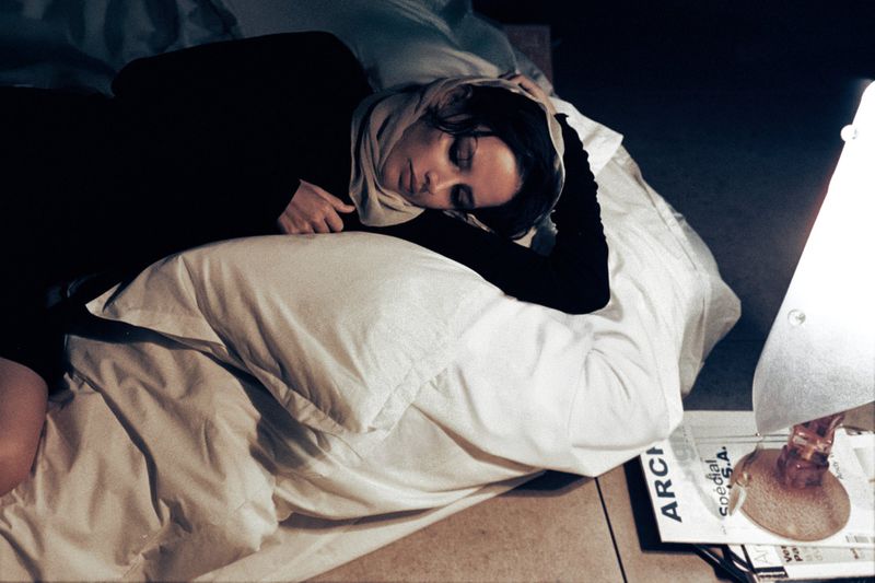 Woman sleeping on a bed with a headscarf on her head, next to a lamp and magazines. She is resting peacefully indoors at night.
