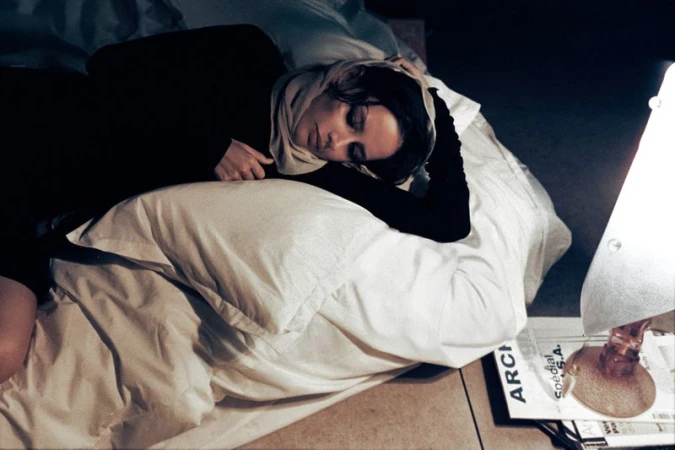 Woman sleeping on a bed with a headscarf on her head, next to a lamp and magazines. She is resting peacefully indoors at night.