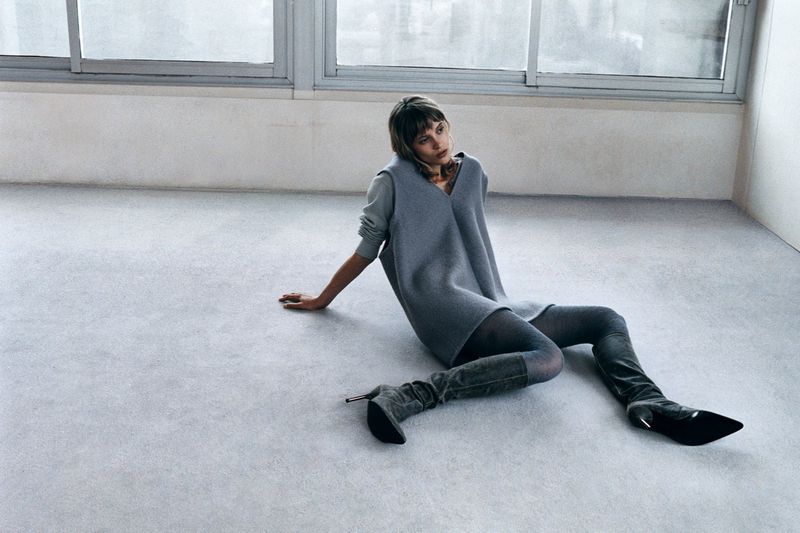 An adult female model in a grey dress, tights, and heeled boots sits in a photo studio with white walls and grey floor near a window.