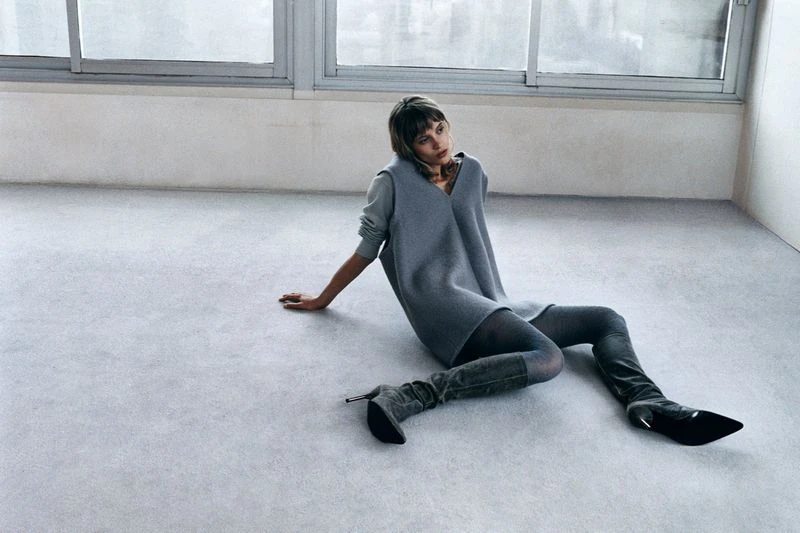 An adult female model in a grey dress, tights, and heeled boots sits in a photo studio with white walls and grey floor near a window.