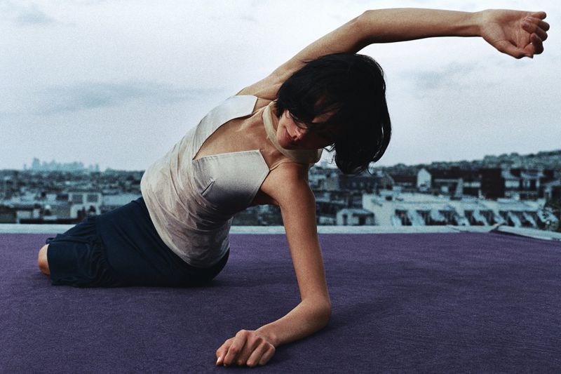Adult female performs yoga on a rooftop, wearing a light-colored top and dark pants. An urban skyline appears in the overcast background.