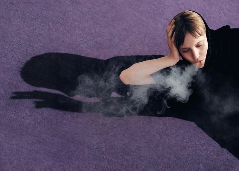 Woman exhales a stream of smoke while lying on a lilac surface, her dark shadow cast on the background with vaporous smoke.