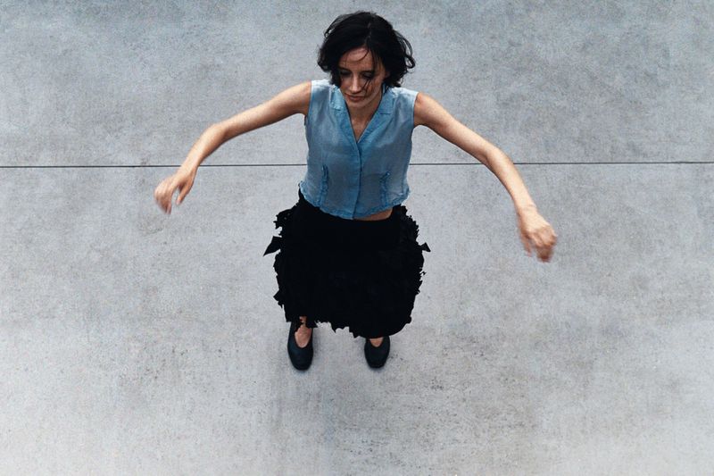 Overhead shot of a woman with dark hair wearing a blue top and black skirt with flat shoes standing on a grey concrete floor with arms out.