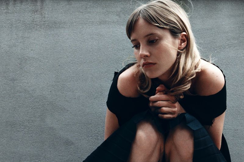 Blonde woman with dark off-the-shoulder dress sits against gray wall. Hands clasped, gazing downward in a close-up shot.