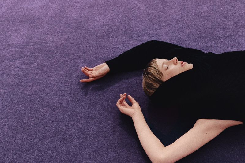 Woman in black sweater lying on a purple textured surface with eyes closed and relaxed posture, creating a serene and minimalist scene.