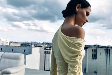 Model seen from behind wearing a draped pale yellow off-the-shoulder top against an urban cityscape from a rooftop.