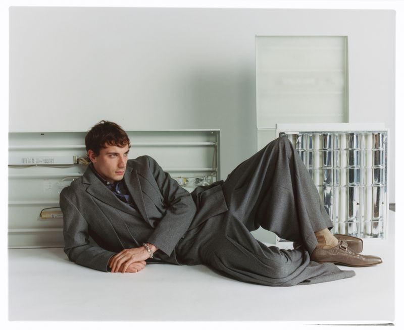 Man in gray suit lies on white surface with fluorescent lights. He is wearing brown loafers, and looking to his left in an indoor setting.