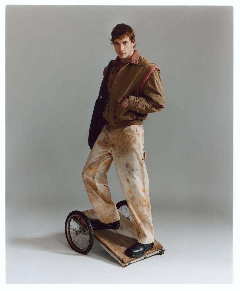 Adult male stands on a wheeled platform in a studio wearing a brown bomber jacket and beige pants with paint splatters against grey backdrop.