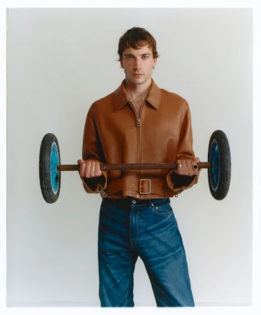 Adult man in a brown jacket and blue jeans posing with a barbell made from a wooden rod and wheels in front of a neutral background.
