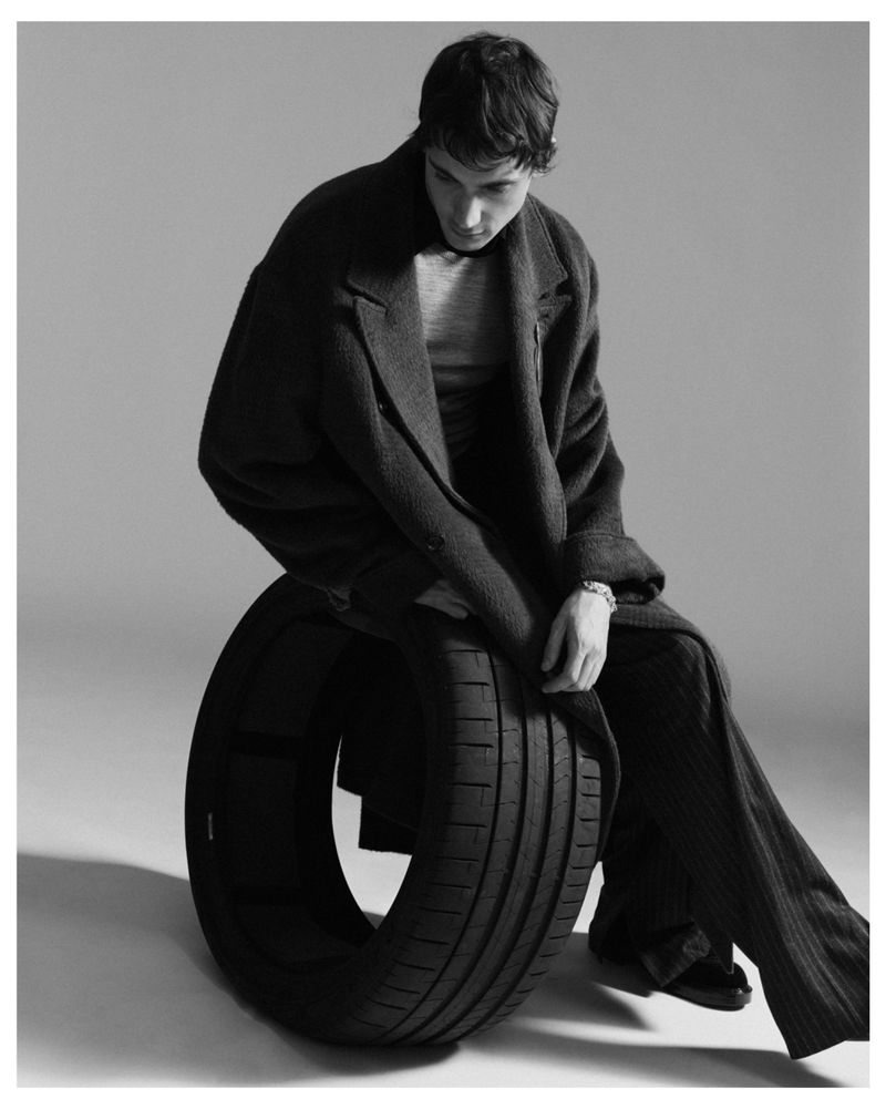 Monochrome studio shot of a young adult man in a coat and pants, sitting on a car tire against a gray backdrop.