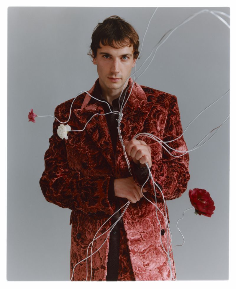 A male model in a burgundy velvet coat poses in a studio setting with floral accents and white wire-like props. Neutral grey background.