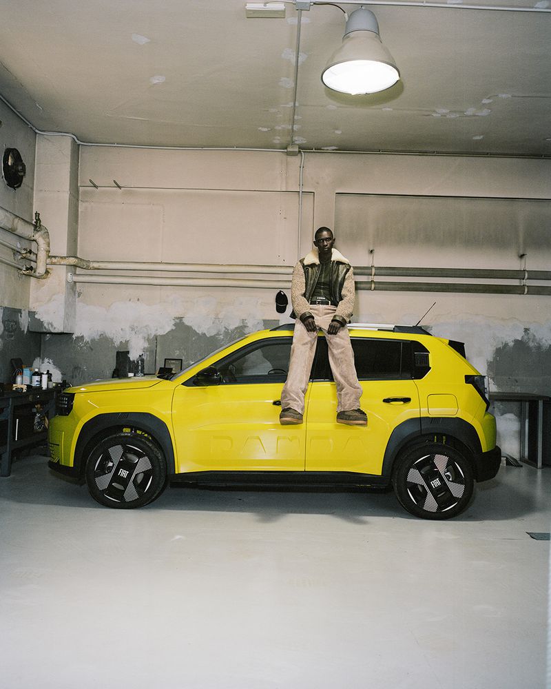 An adult man is sitting on the roof of a yellow SUV in an indoor industrial setting with concrete walls, fluorescent lighting, and workshop equipment.