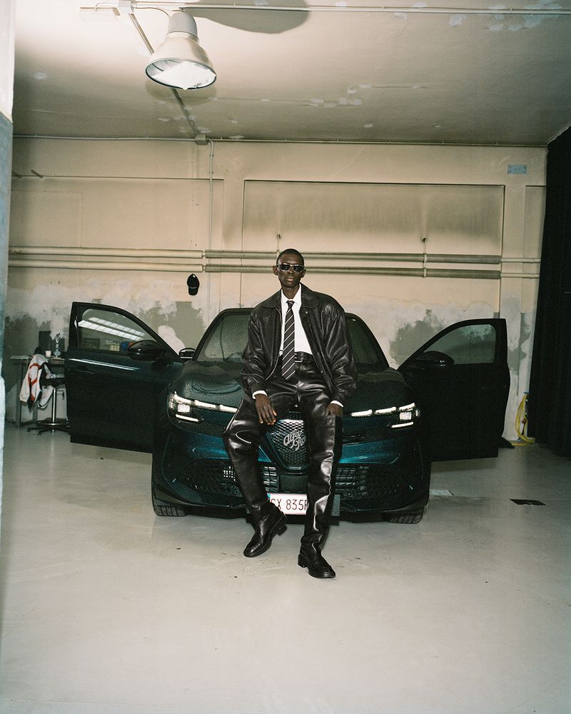 An adult man in leather clothing sits on the hood of a car in an indoor garage setting, illuminated by an overhead light.