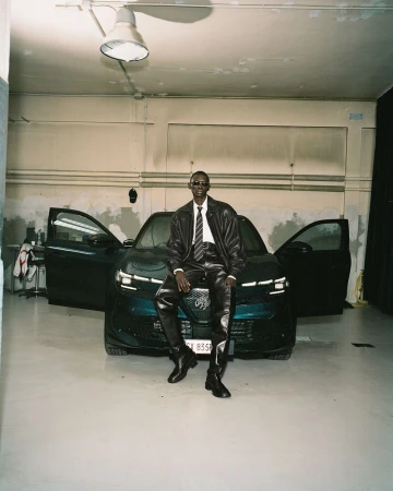 An adult man in leather clothing sits on the hood of a car in an indoor garage setting, illuminated by an overhead light.