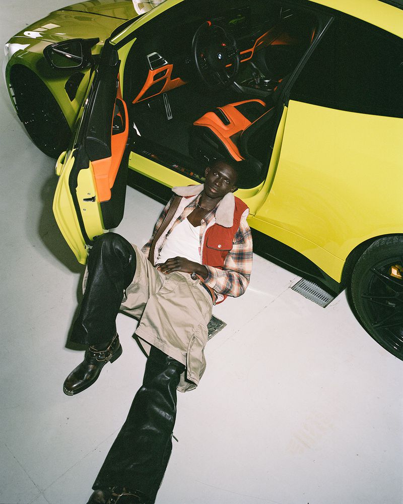 A man in a plaid shirt and vest is lying next to a yellow sports car in a photo studio with black boots and light-colored pants.