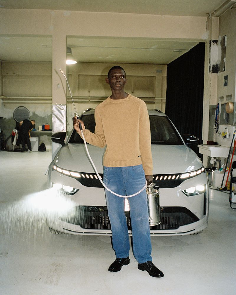 A stylish man poses with a sprayer and a modern white car in a garage, showcasing automotive fashion in an industrial setting.