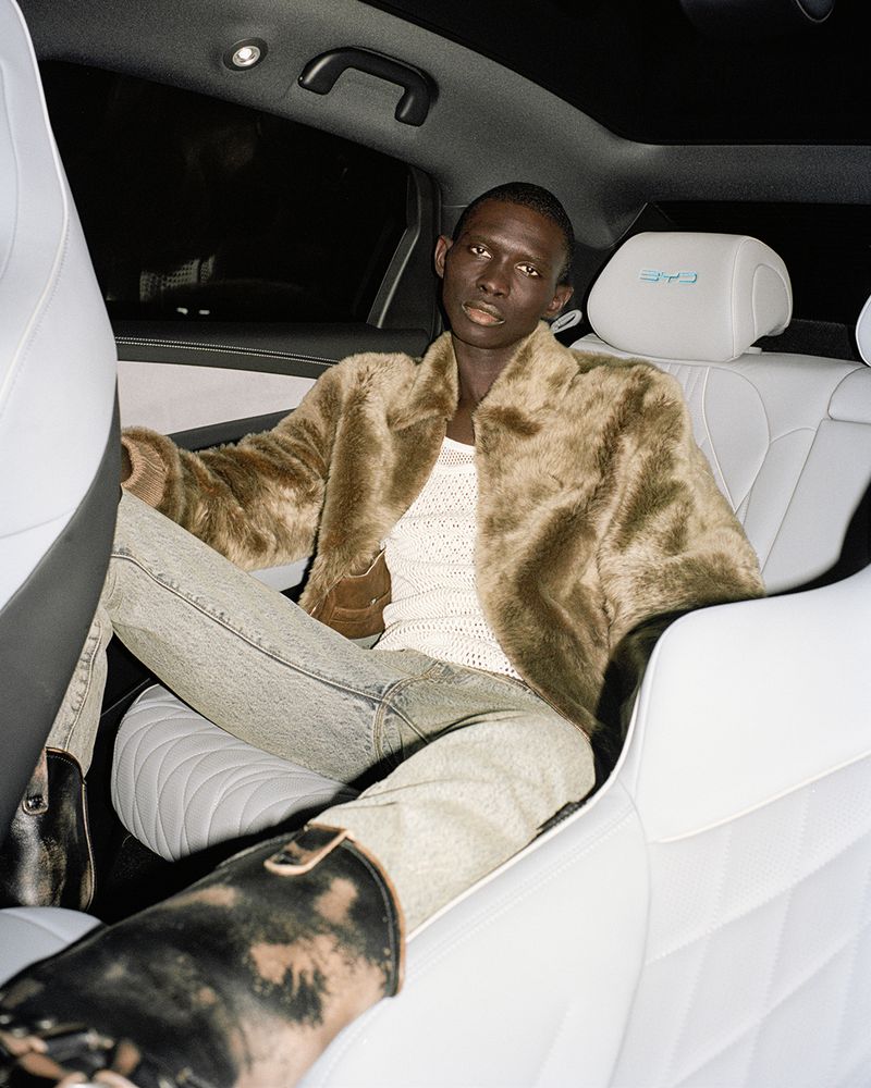 A stylish adult male sits in the back of a luxury car, wearing a fur jacket, light denim jeans, and black boots, against a dark interior.