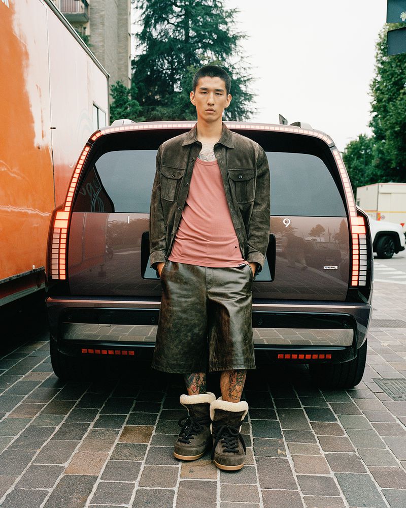 An Asian man stands in front of a modern SUV in an urban setting, wearing a brown jacket, pink shirt, leather shorts, and fur-lined boots.