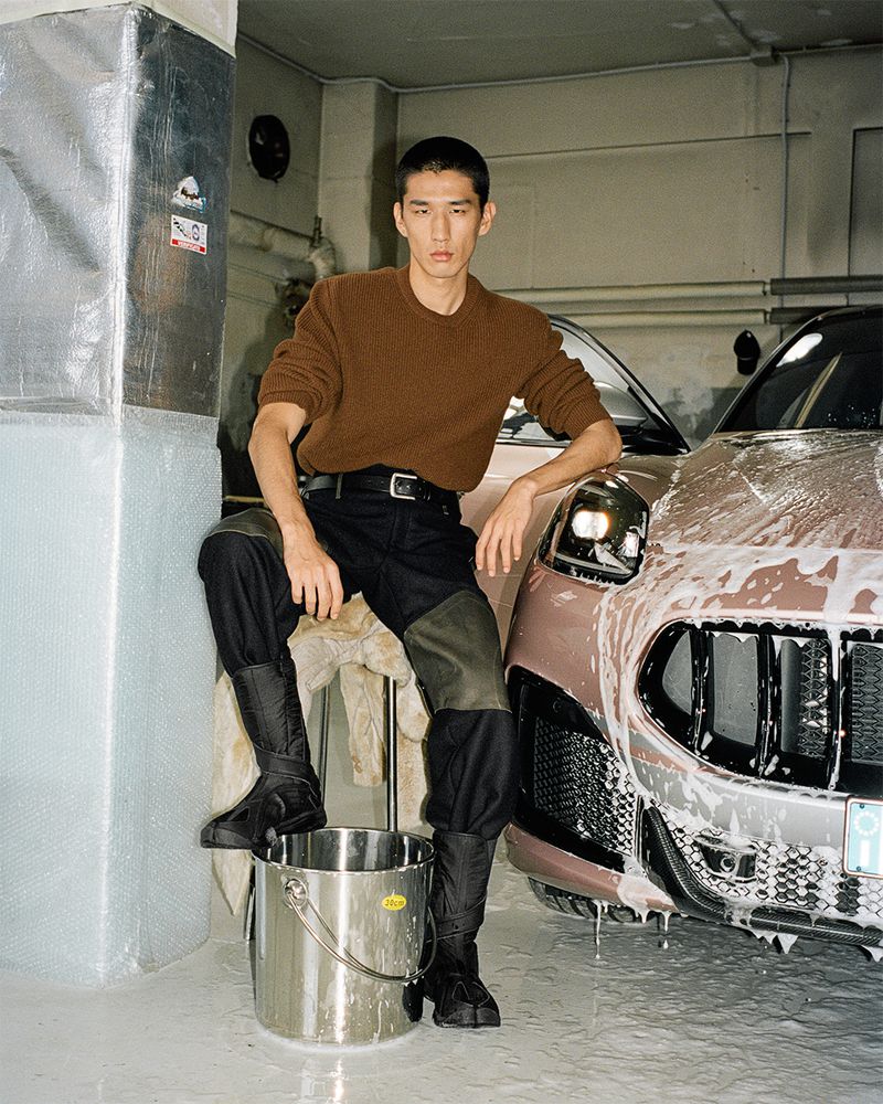 Man poses with a soap-covered SUV inside an indoor garage-like space. He is wearing a brown sweater, black pants, and black boots.