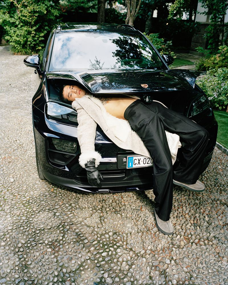 Man in white coat reclines on black Porsche in outdoor setting, featuring cobblestone ground, trees, and sleek car design.
