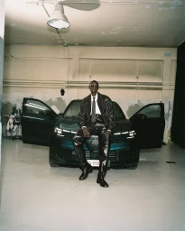 Model Fernando Cabral wearing a black leather suit and sunglasses seated on the hood of a car inside a garage.