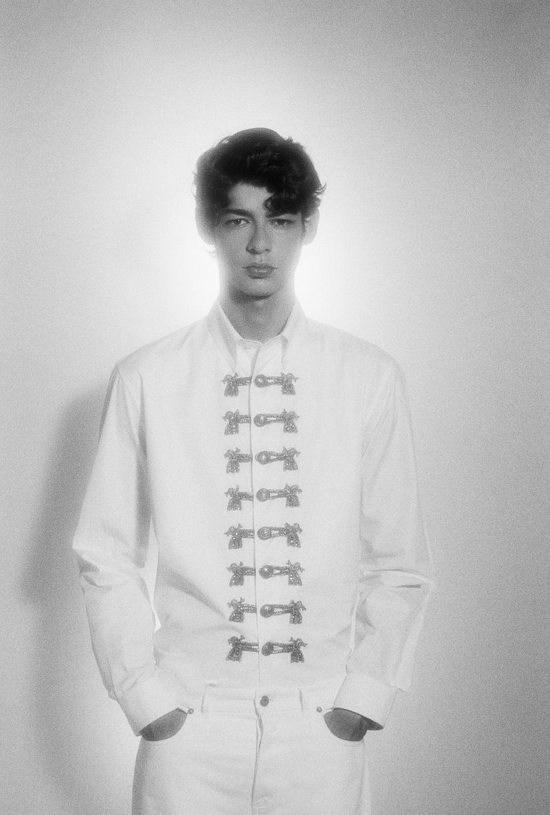 Monochrome portrait of a young adult man in a white button-down shirt with decorative clasps, against a bright, diffused background.