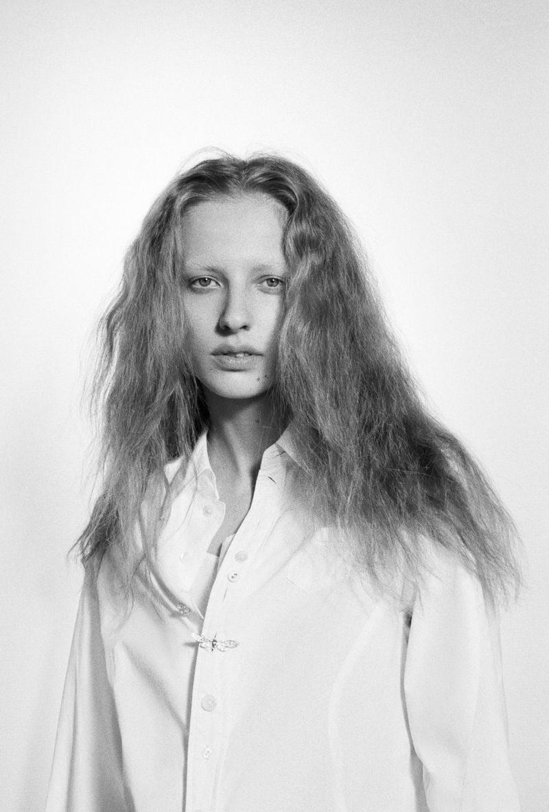 Black and white portrait of a young adult female model with long, wavy hair, wearing a white button-down shirt against a plain background.