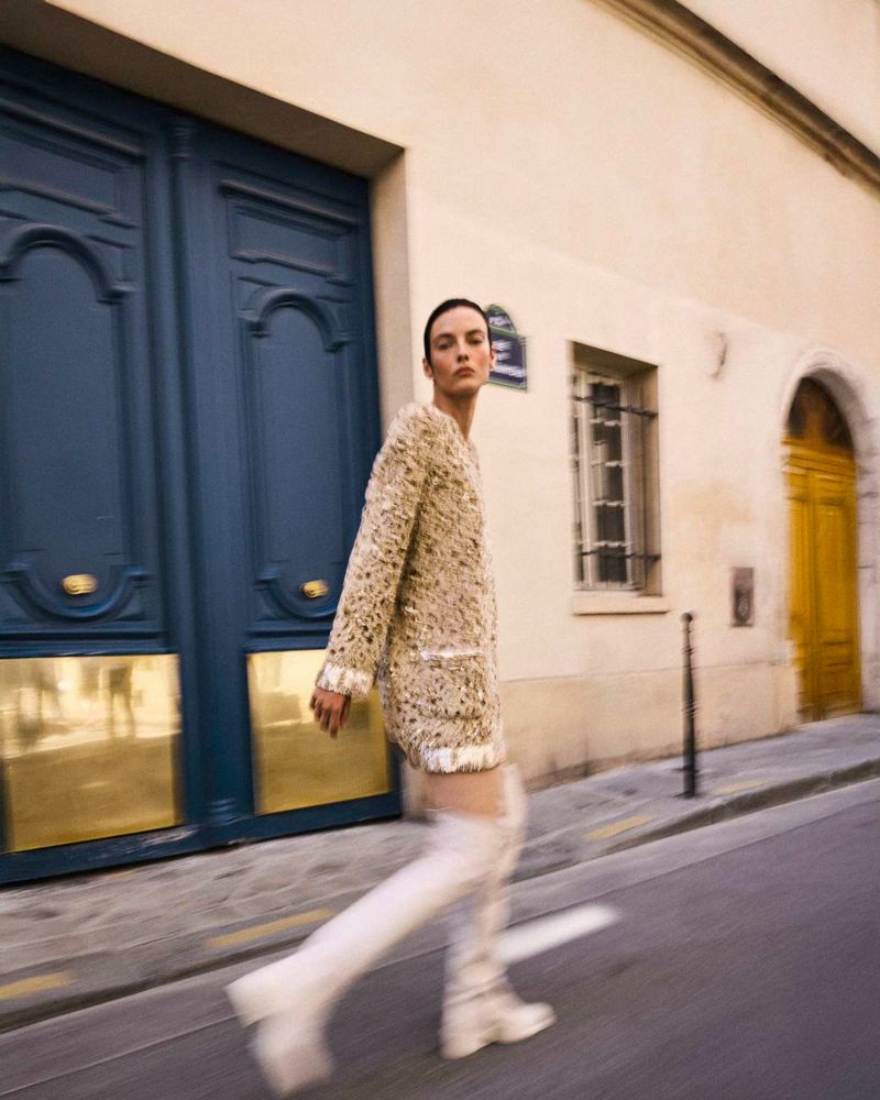 Fashion photography featuring a woman in a textured beige dress and white boots, walking on a city street in front of blue doors.