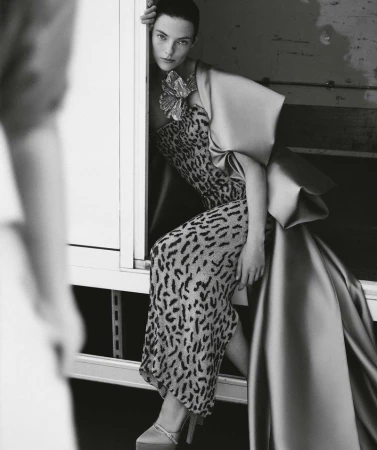 A woman poses elegantly in a leopard-print dress with a large satin bow and platform heels, positioned on a white surface, in black and white.