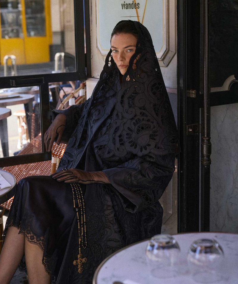 Woman in black lace dress and head covering sitting at an outdoor cafe with glasses on the table, creating a moody fashion shot.